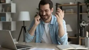 Smiling man at home office next to laptop speaking on smartphone. VoIP phone, cellphone service