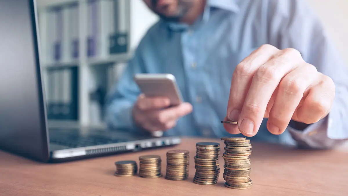 Man counting coins next to laptop while holding smart phone.