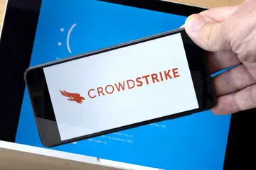 hand holding phone with CrowdStrke Logo in front of a PC with a blue screen.